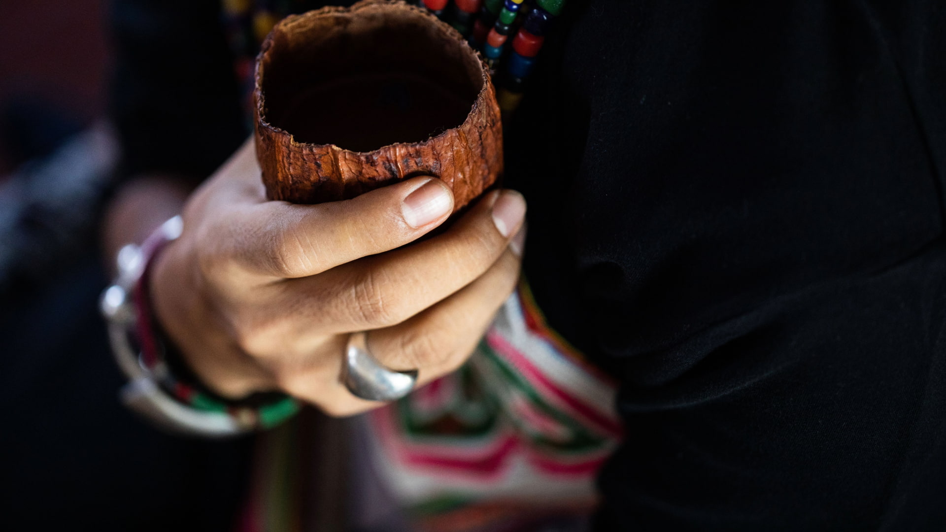 Hand holding a ceremonial cup during a spiritual ritual, with traditional jewelry and colorful attire.