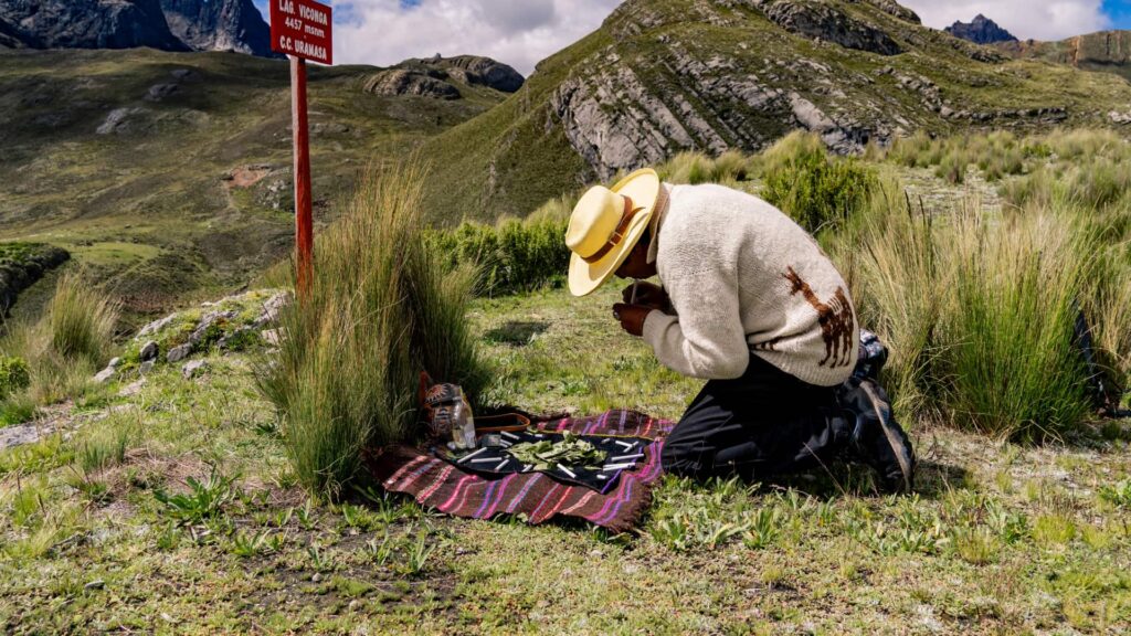 Man performing a traditional Andean offering ceremony with coca leaves in the mountains, kneeling on a colorful textile.