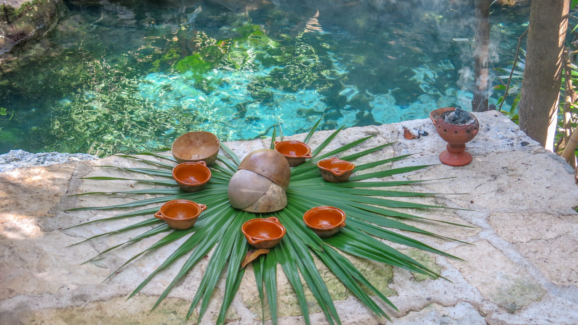 Ceremonial items arranged on a palm leaf near a sacred water pool, used in a traditional healing ritual with bowls and incense.