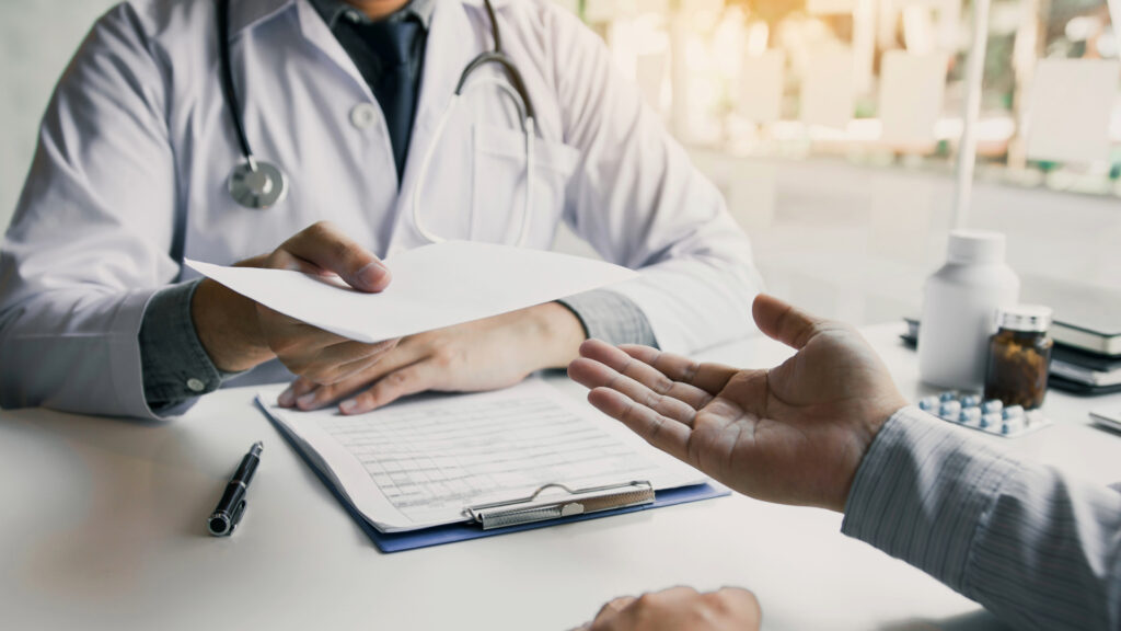 Doctor handing a prescription to a patient across a desk during a medical consultation, with a clipboard and medical items in the background.