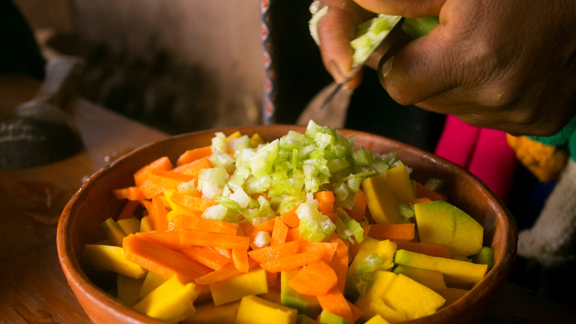 Bowl of freshly chopped vegetables, including carrots, squash, and onions, with a hand adding more ingredients.