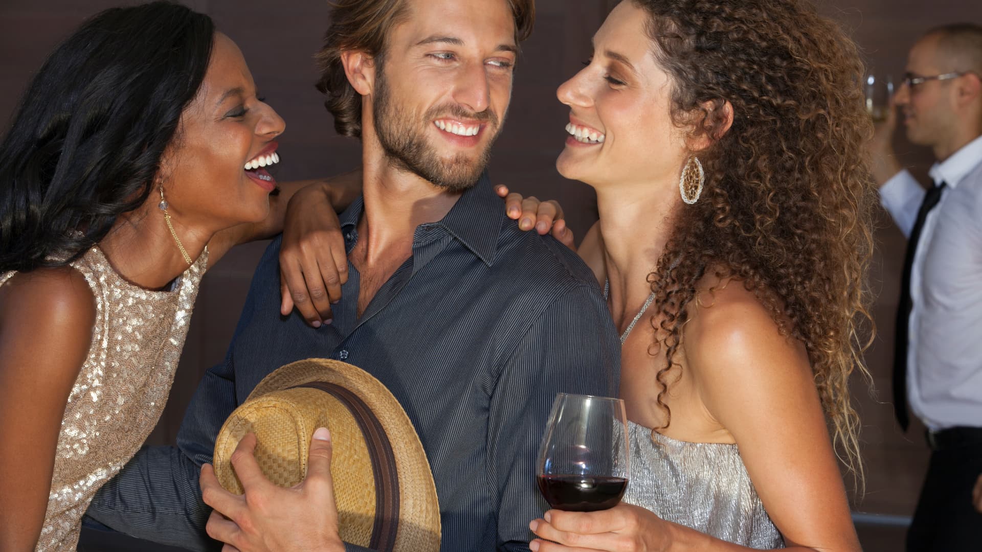 Three friends, two women and a man, laughing and embracing while enjoying a social gathering, with one of them holding a glass of wine.