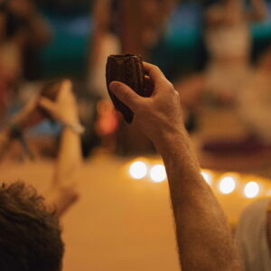 A participant’s hand holding ayahuasca during a group spiritual ceremony, with people in the background and lit candles.