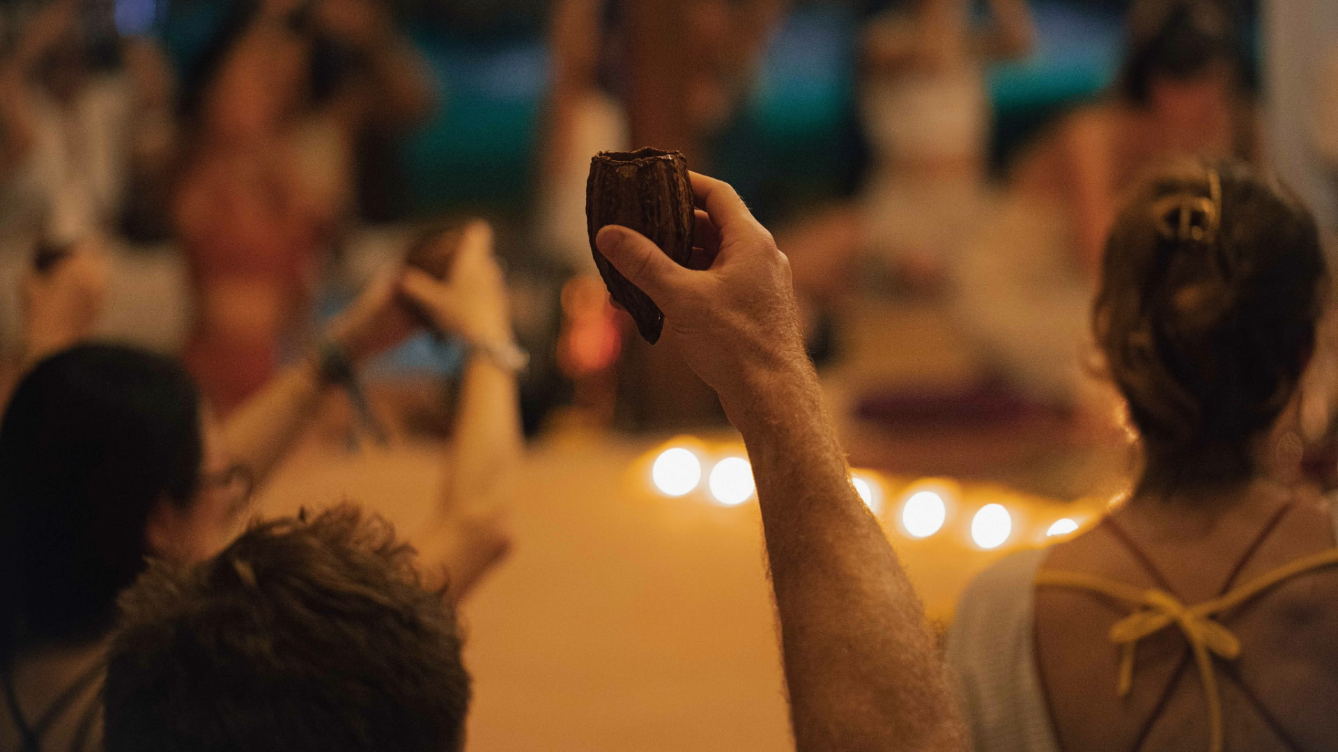 A participant’s hand holding ayahuasca during a group spiritual ceremony, with people in the background and lit candles.