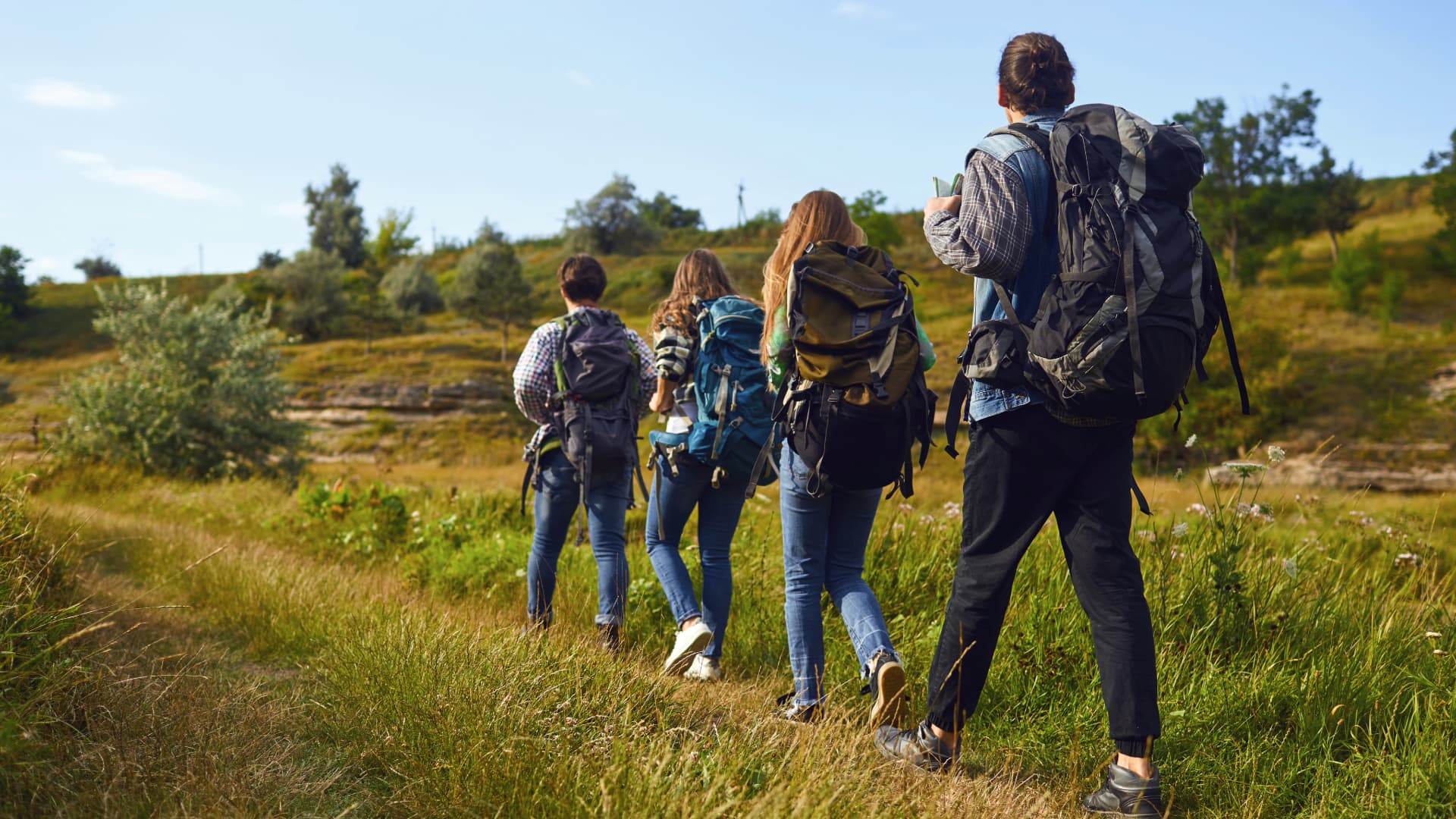 Four hikers with backpacks walking along a scenic trail in a grassy, open landscape with trees and hills in the background.