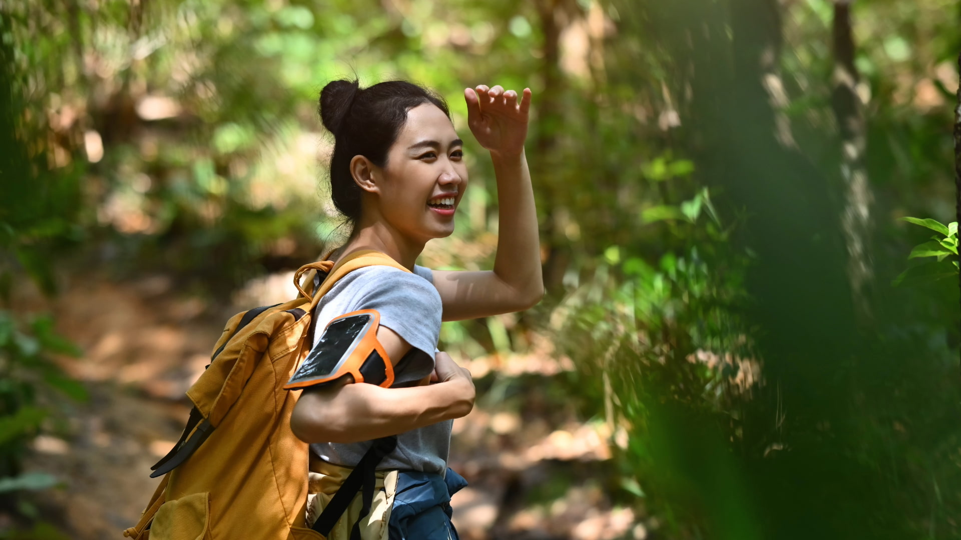 Smiling woman with a backpack on a forest trail, raising her hand to shield her eyes from the sunlight while hiking.