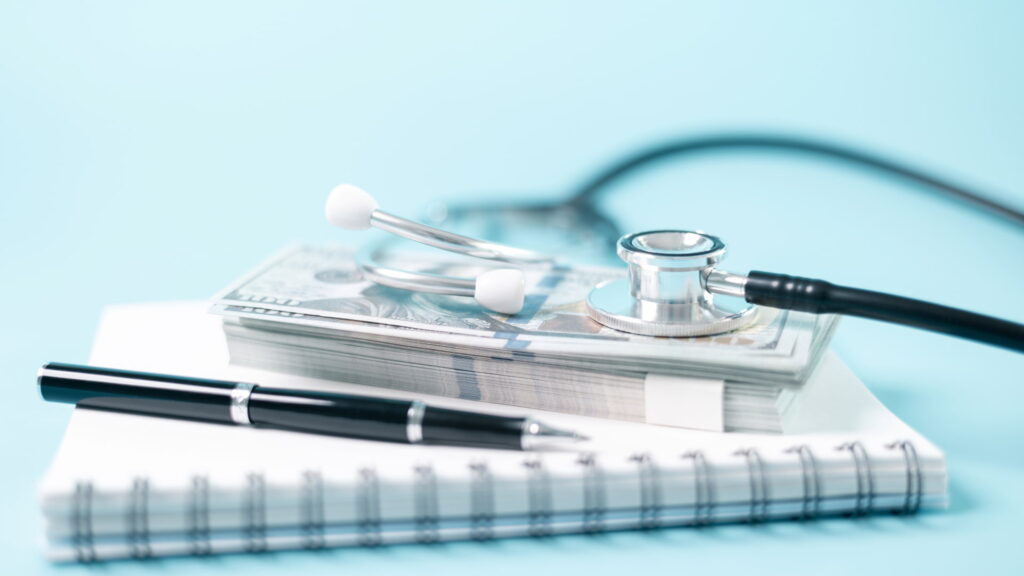 Stethoscope on top of a stack of dollar bills with a pen and notebook, representing healthcare costs and financial planning.