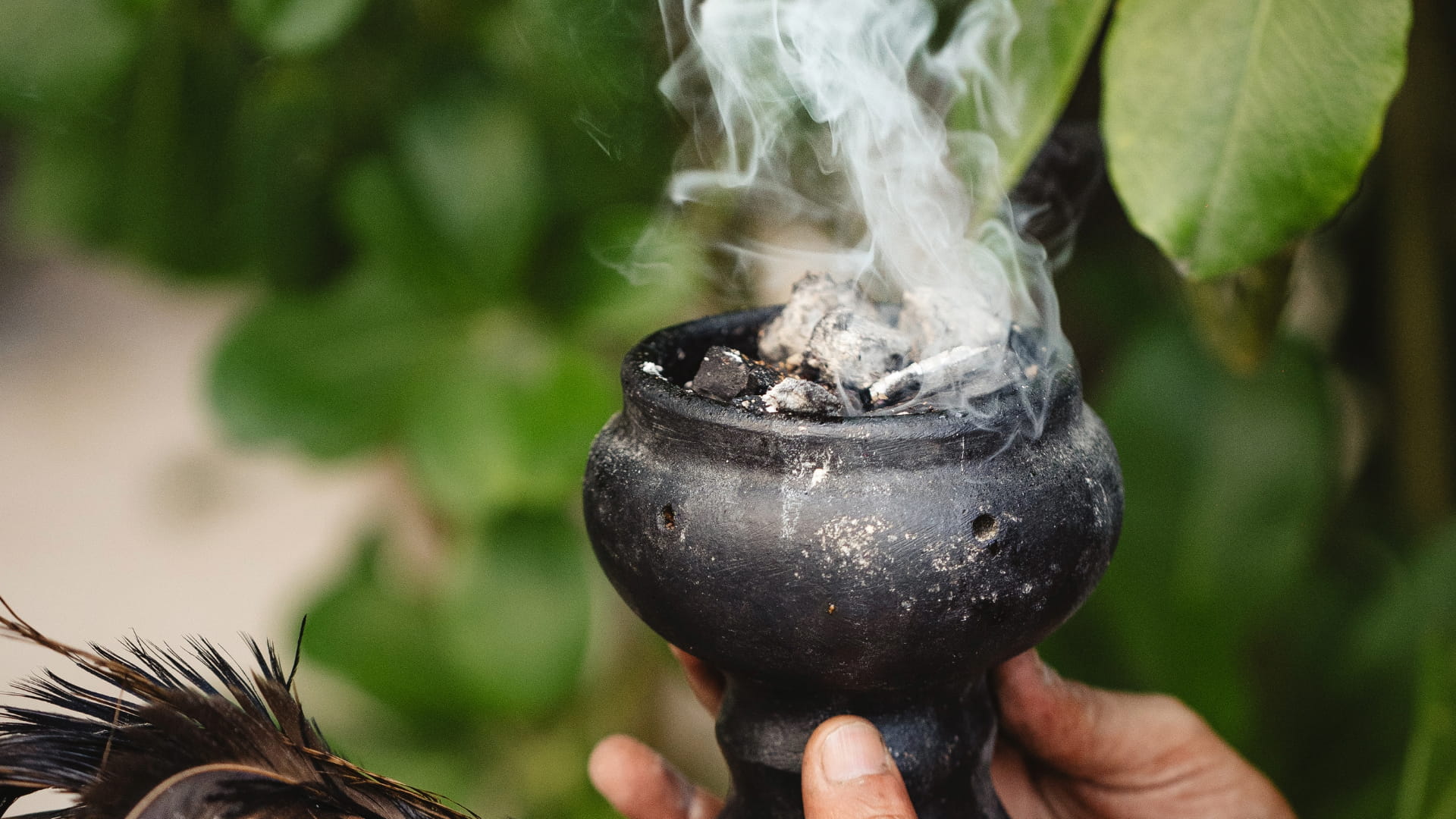 Hand holding a smoking incense burner during a cleansing ritual, with smoke rising against a natural green background.