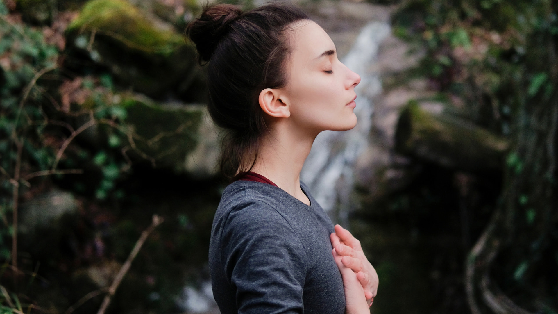 Woman with closed eyes and hand on her chest, meditating peacefully in a natural outdoor setting.