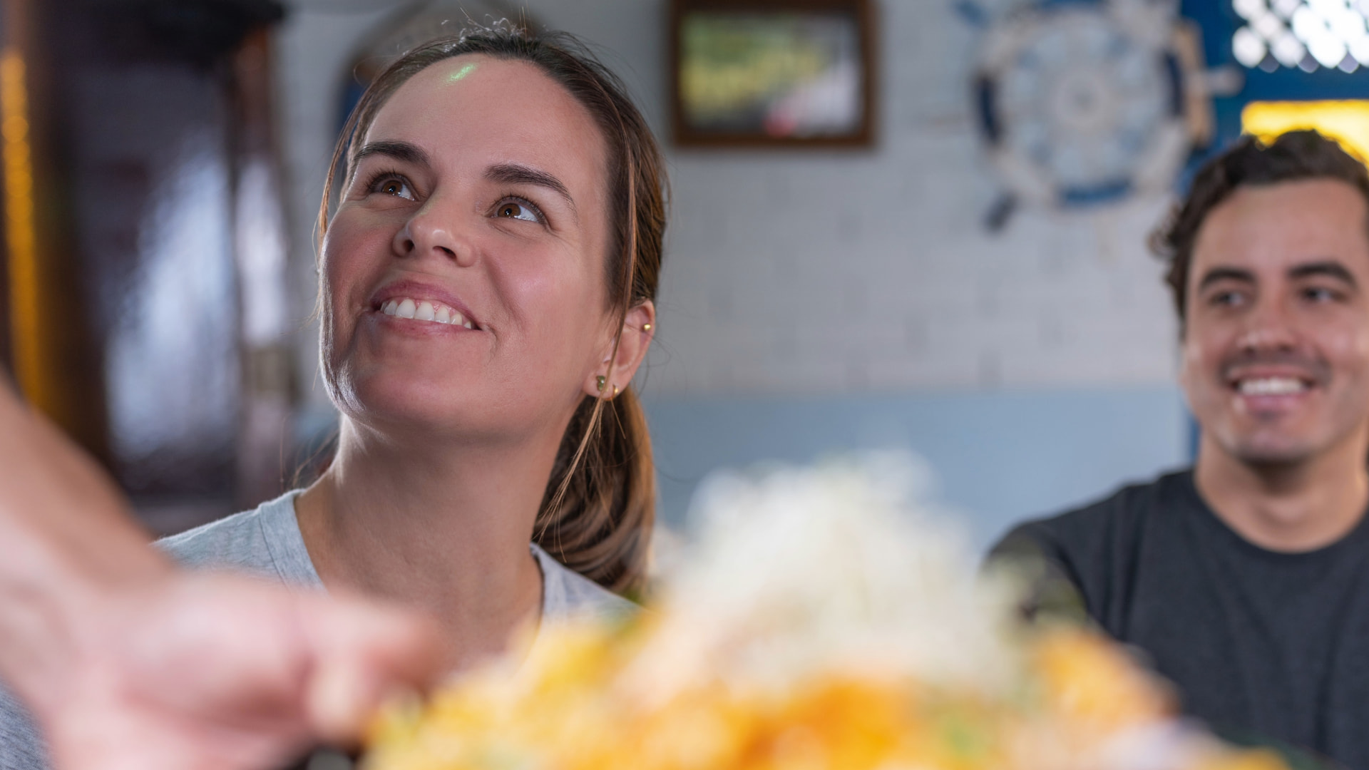 Smiling woman looking up, seated at a restaurant table with a plate of food in front, enjoying a pleasant dining experience.