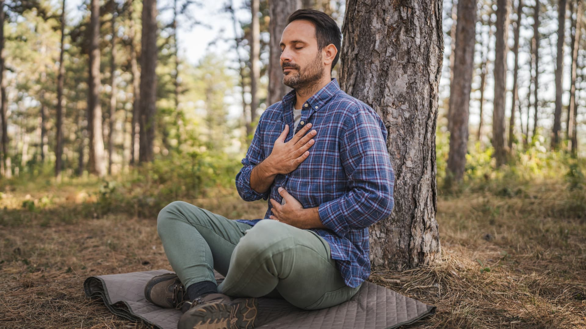 Man sitting on a mat in the forest, with eyes closed and hands on his chest and stomach, engaging in deep breathing and meditation for relaxation and connection with nature.