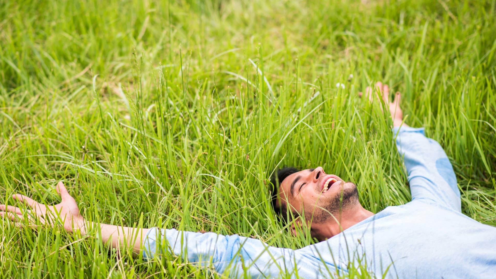 Man lying in a grassy field with a joyful expression, arms outstretched, embracing relaxation and connection with nature.