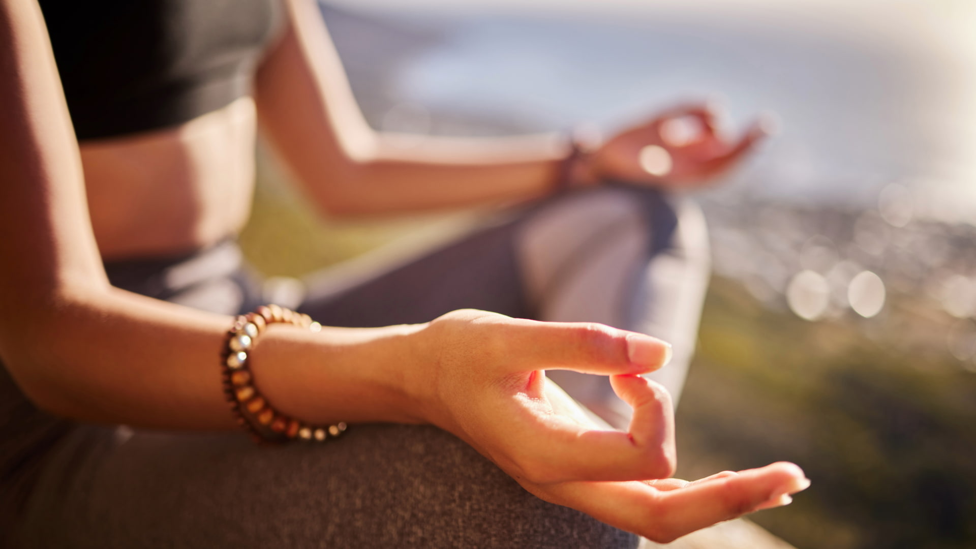 Close-up of hands in a meditative mudra position while a person practices meditation outdoors with a blurred natural background.