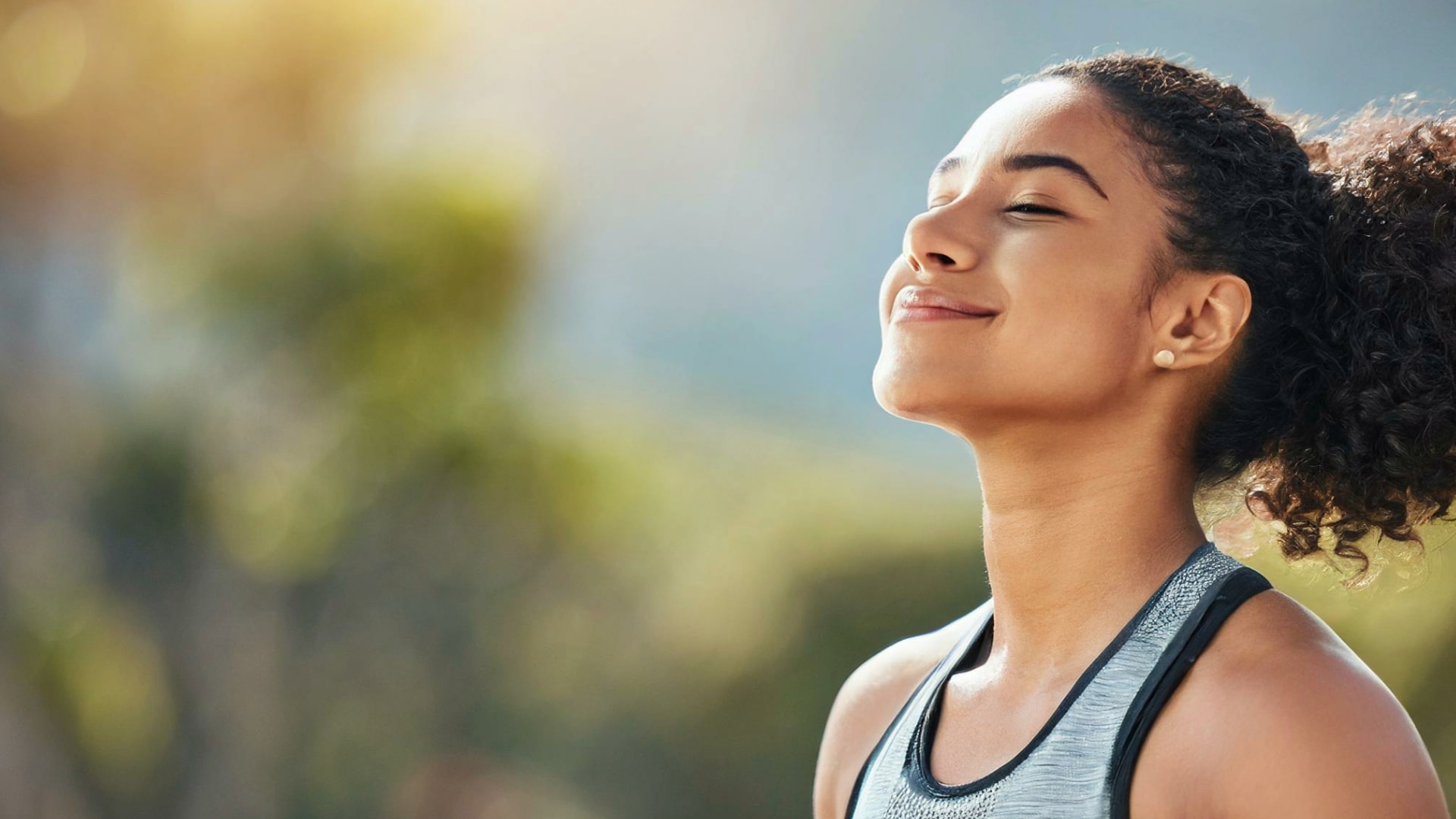 Young woman with closed eyes and a relaxed smile, experiencing mindfulness outdoors with a blurred natural background.