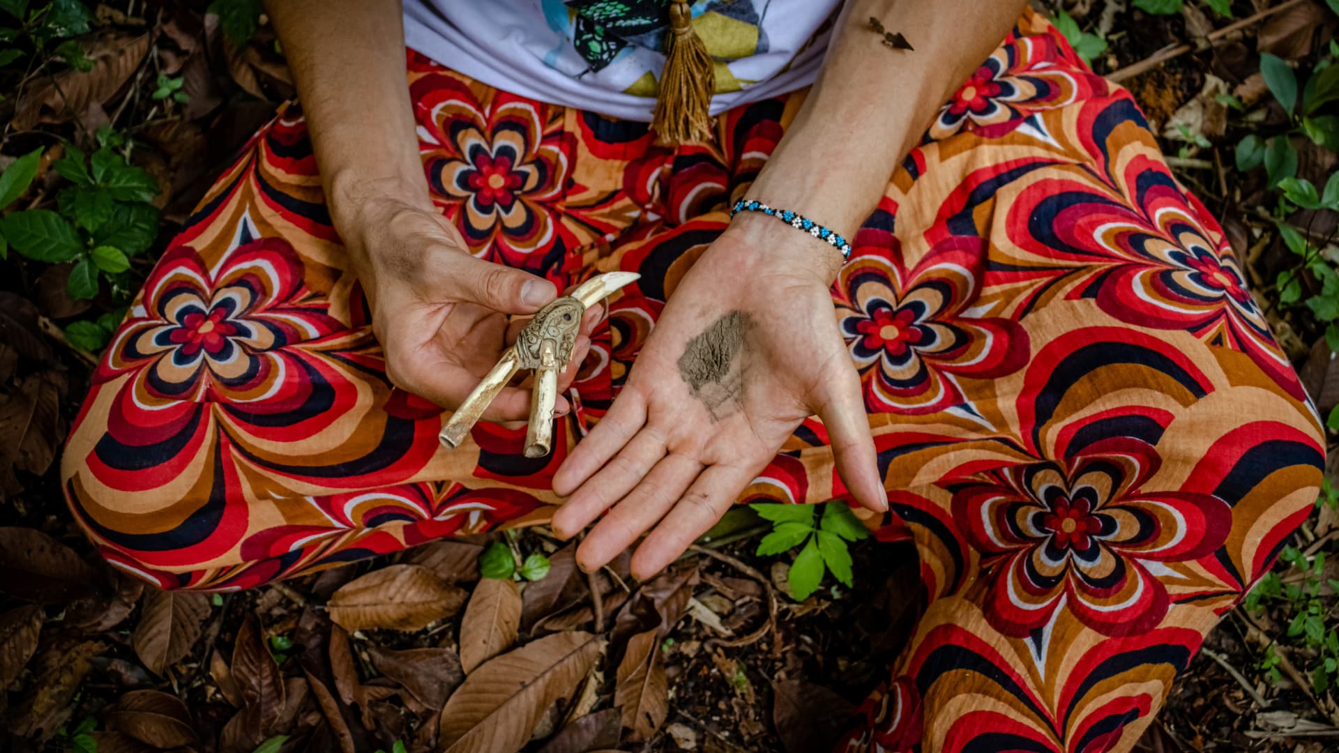 Person seated outdoors in vibrant patterned clothing, holding a sacred object in one hand and showing ash in the other, representing a moment of ritual and connection with nature.