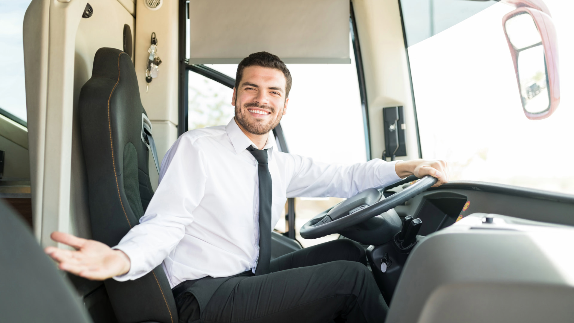 Smiling bus driver in a white shirt and black tie, sitting in the driver's seat and welcoming passengers onboard.