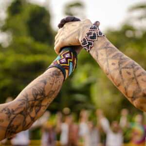 Two tattooed hands joined in a gesture of connection, adorned with colorful traditional woven bracelets, set against a natural background with trees.