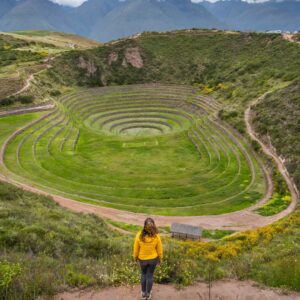 Woman in a yellow jacket admiring the circular terraces of Moray, a historic Inca site with green, layered earth formations set against mountainous terrain.