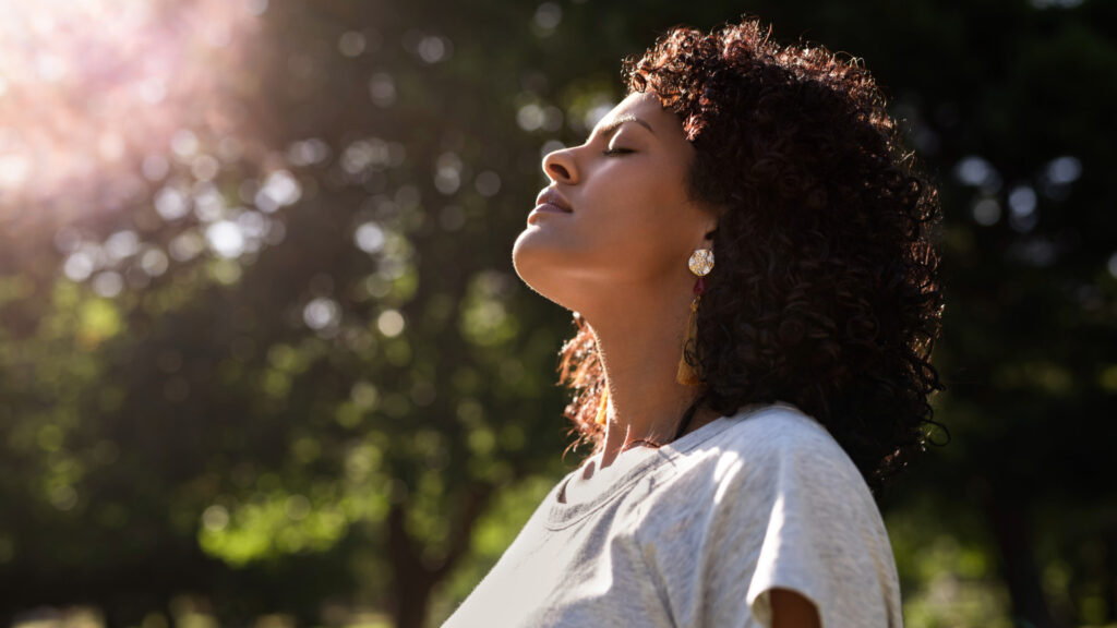 Woman with curly hair and earrings, standing outdoors with eyes closed, basking in the sunlight, surrounded by nature.