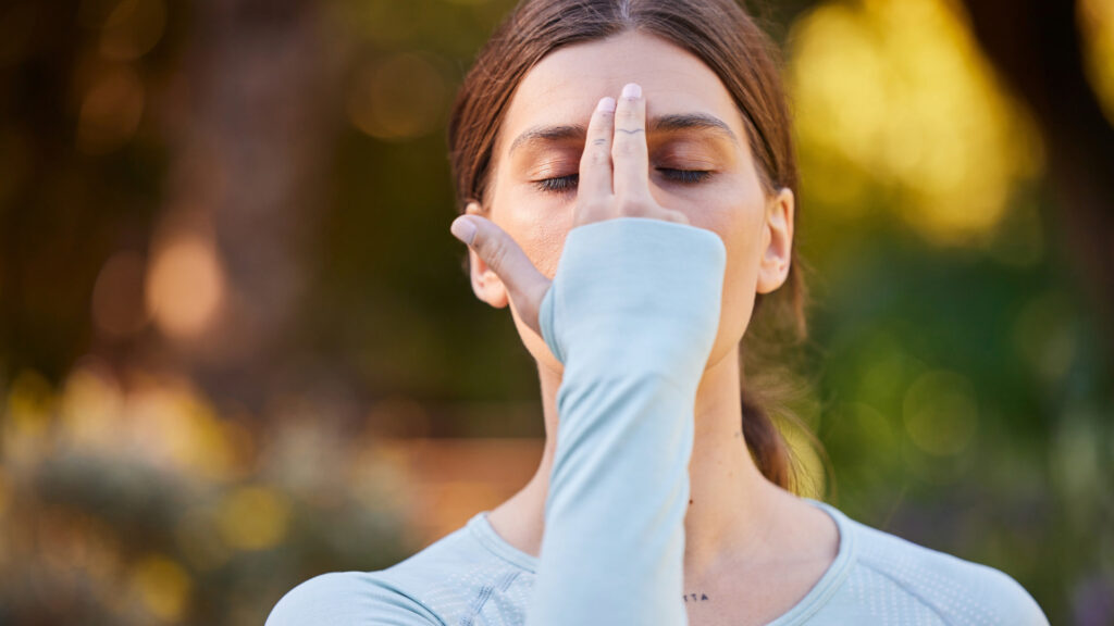 Woman practicing a breathing exercise with closed eyes, using her hand to close one nostril, enhancing mindfulness and relaxation outdoors.